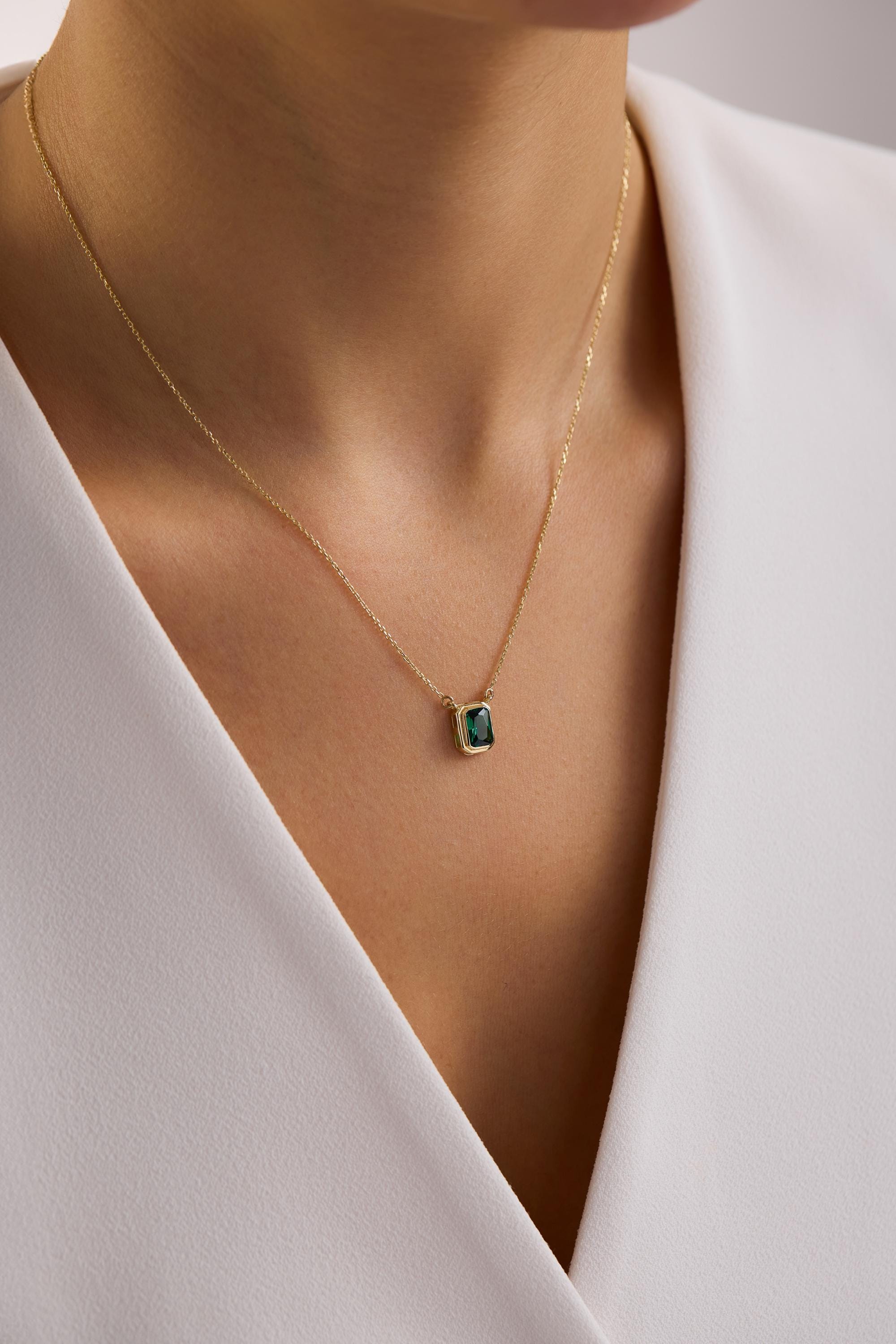 May include: A gold necklace featuring a square, emerald-cut, dark green gemstone pendant. The pendant is set in a gold bezel and hangs from a delicate gold chain. The necklace is displayed against a white background.