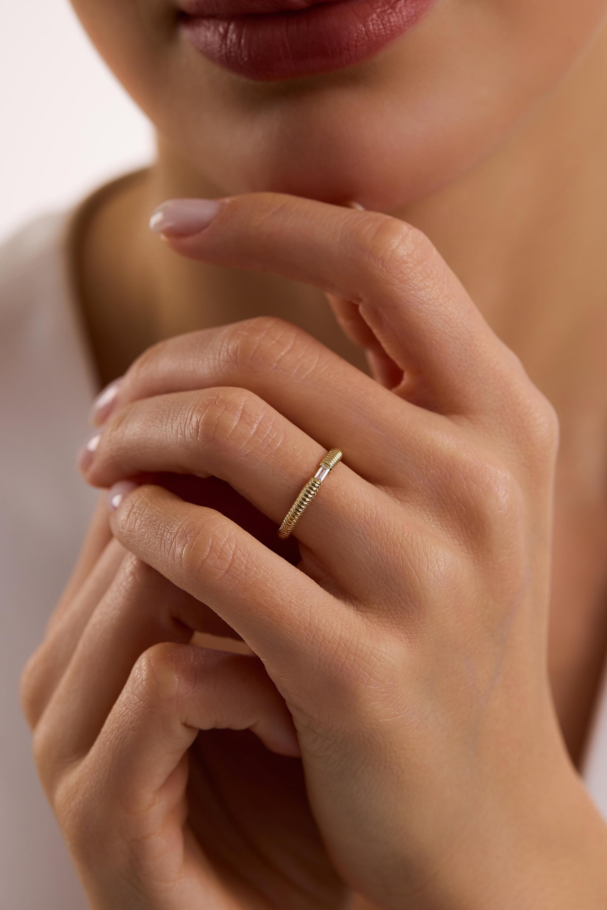 May include: A gold ring with a textured band and a small, rectangular, clear stone. The ring is worn on a finger, showcasing its delicate design and elegant simplicity. The ring's band has a ribbed texture.