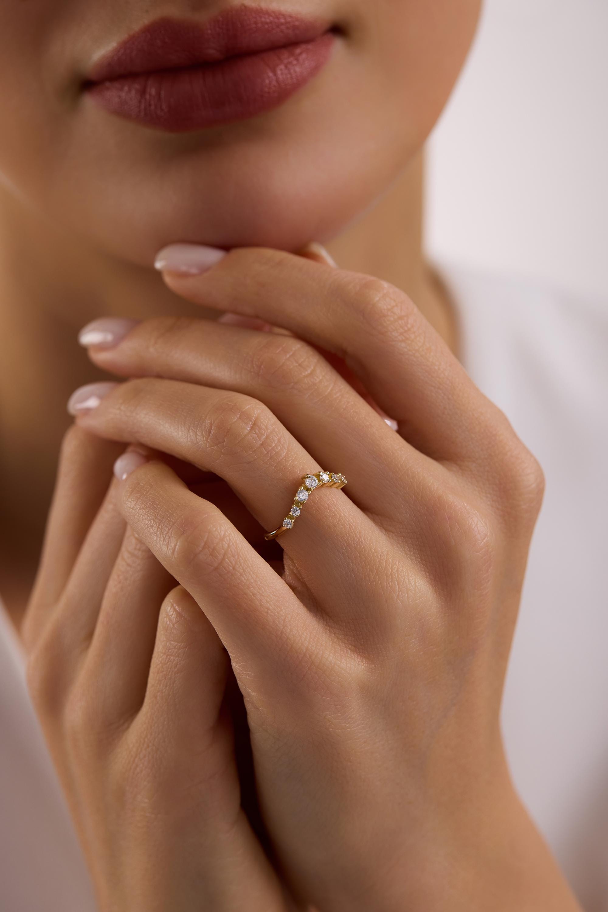 May include: A gold-colored ring with a curved design, set with multiple small, clear gemstones. The ring is worn on a finger, with the hand resting near the face. The ring's design is a delicate arc, with the gemstones adding sparkle.