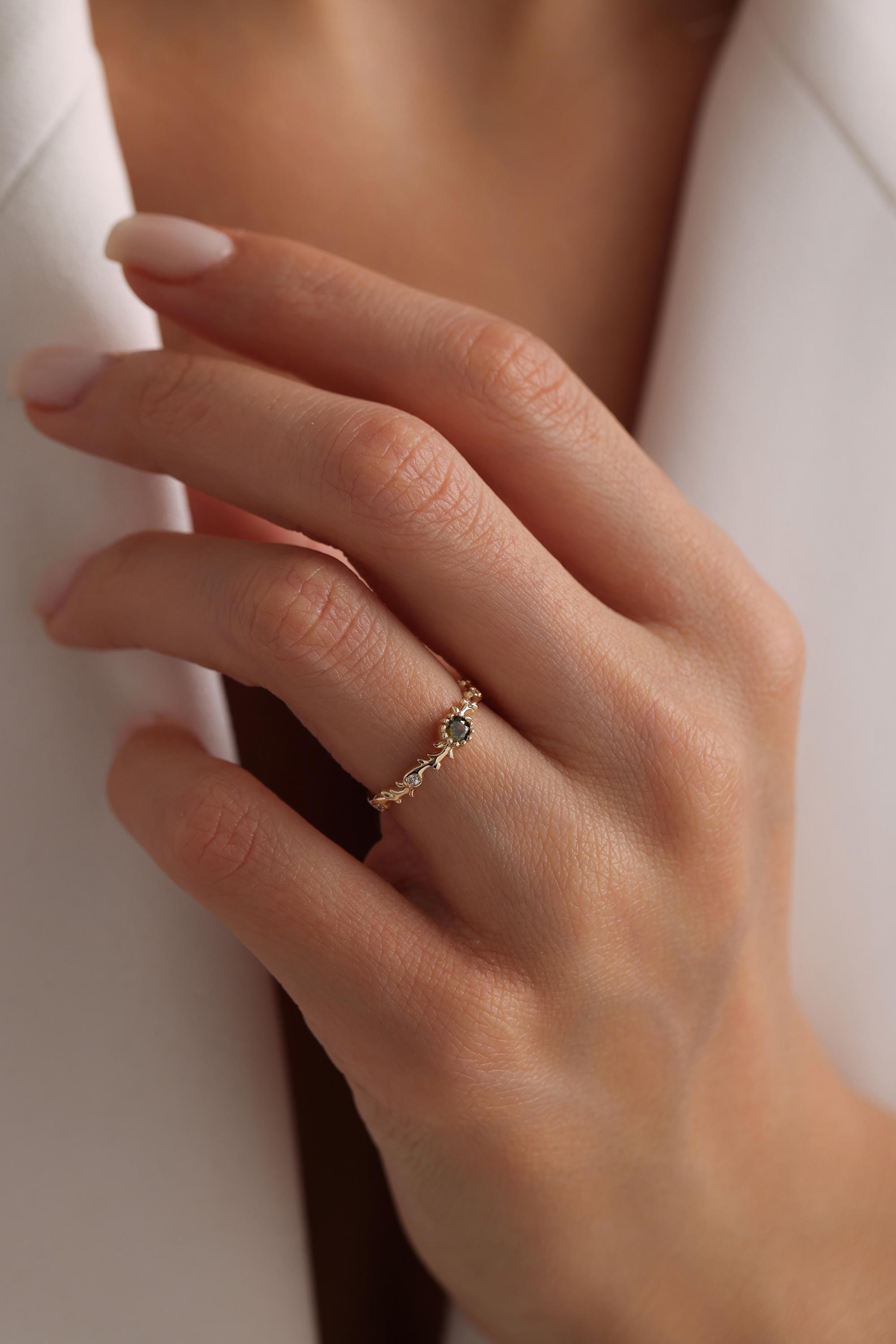 May include: A gold ring with a delicate design featuring a small, dark gray gemstone in the center. The ring is set with smaller, clear gemstones around the center stone.