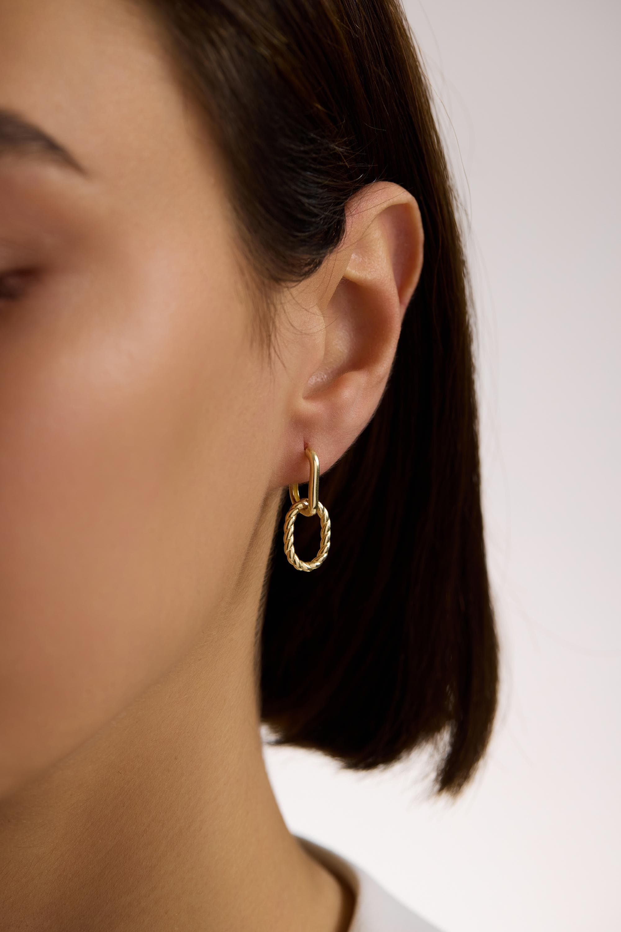 May include: Gold-toned hoop earrings with a textured oval pendant. The earrings are connected by a rectangular link. The oval pendant has a twisted rope design. The earrings are worn on the ear.