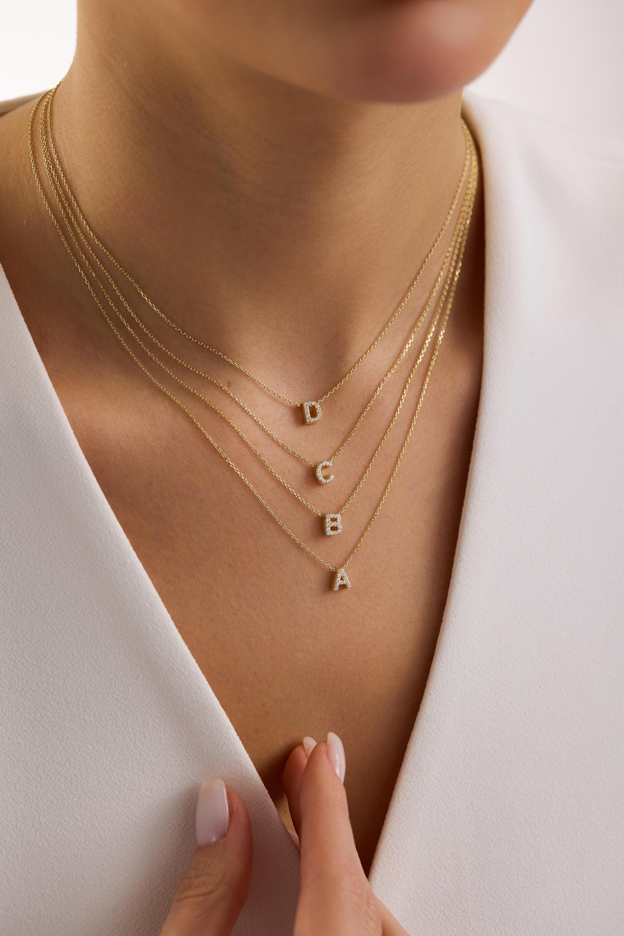 May include: A collection of four delicate gold necklaces, each with a different initial pendant: A, B, C, and D. The pendants are encrusted with small, sparkling stones. The necklaces are layered, creating a stylish and personalized look.