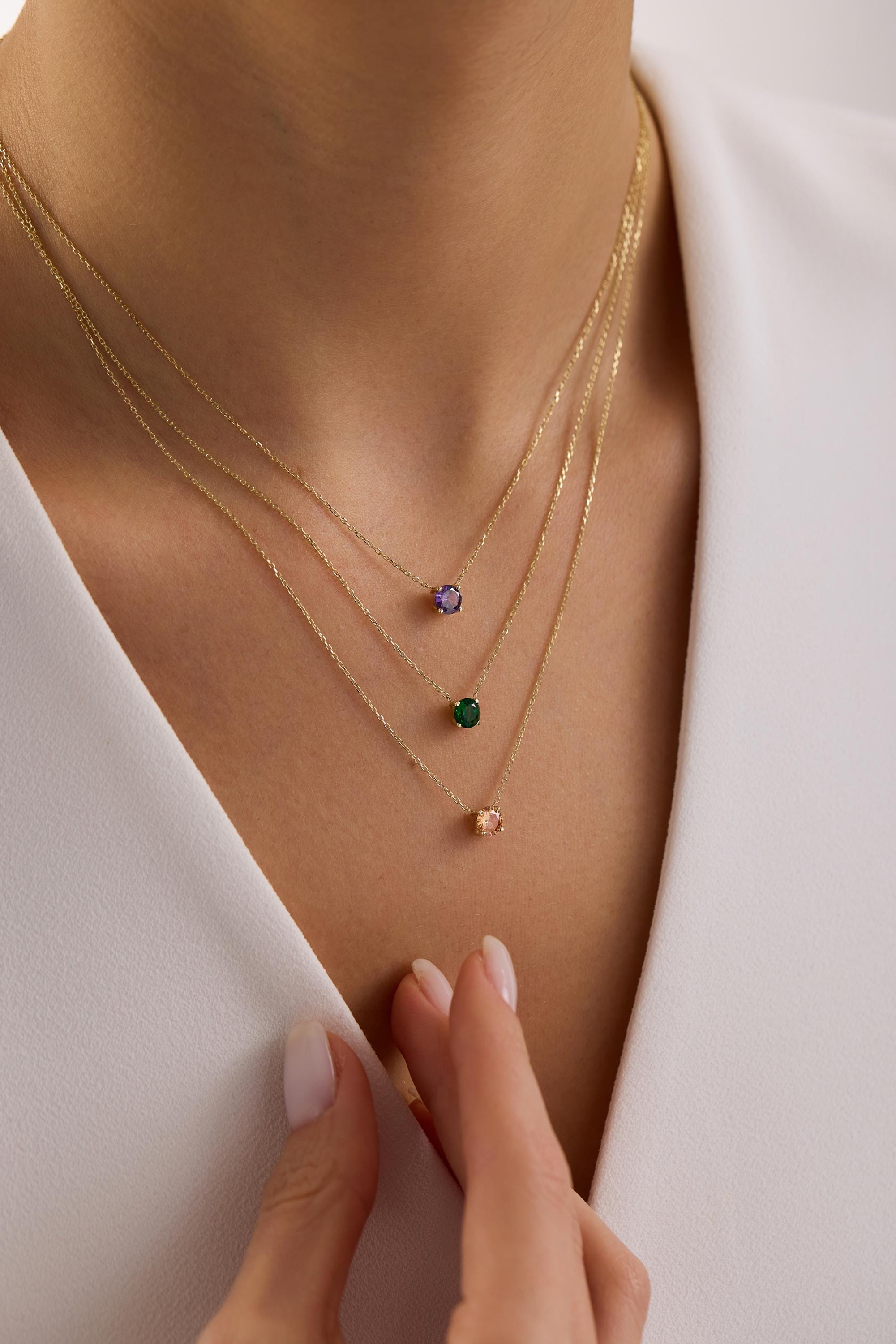 May include: Three delicate gold necklaces, each featuring a single, round gemstone pendant. The gemstones are purple, green, and peach-colored. The necklaces are layered and worn against a white top.